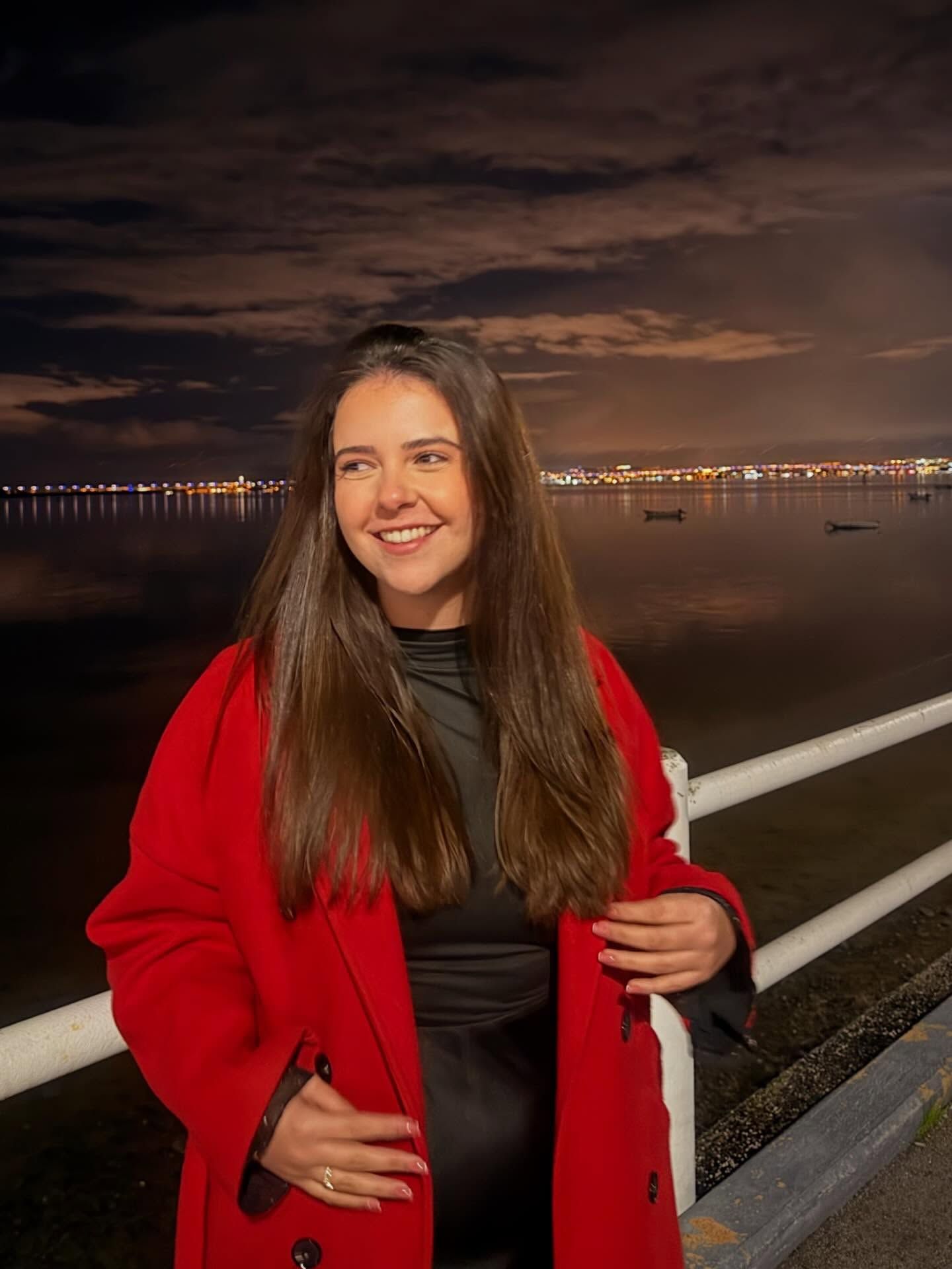 Carolina Mendonça, de casaco vermelho, a sorrir junto ao rio ao anoitecer.