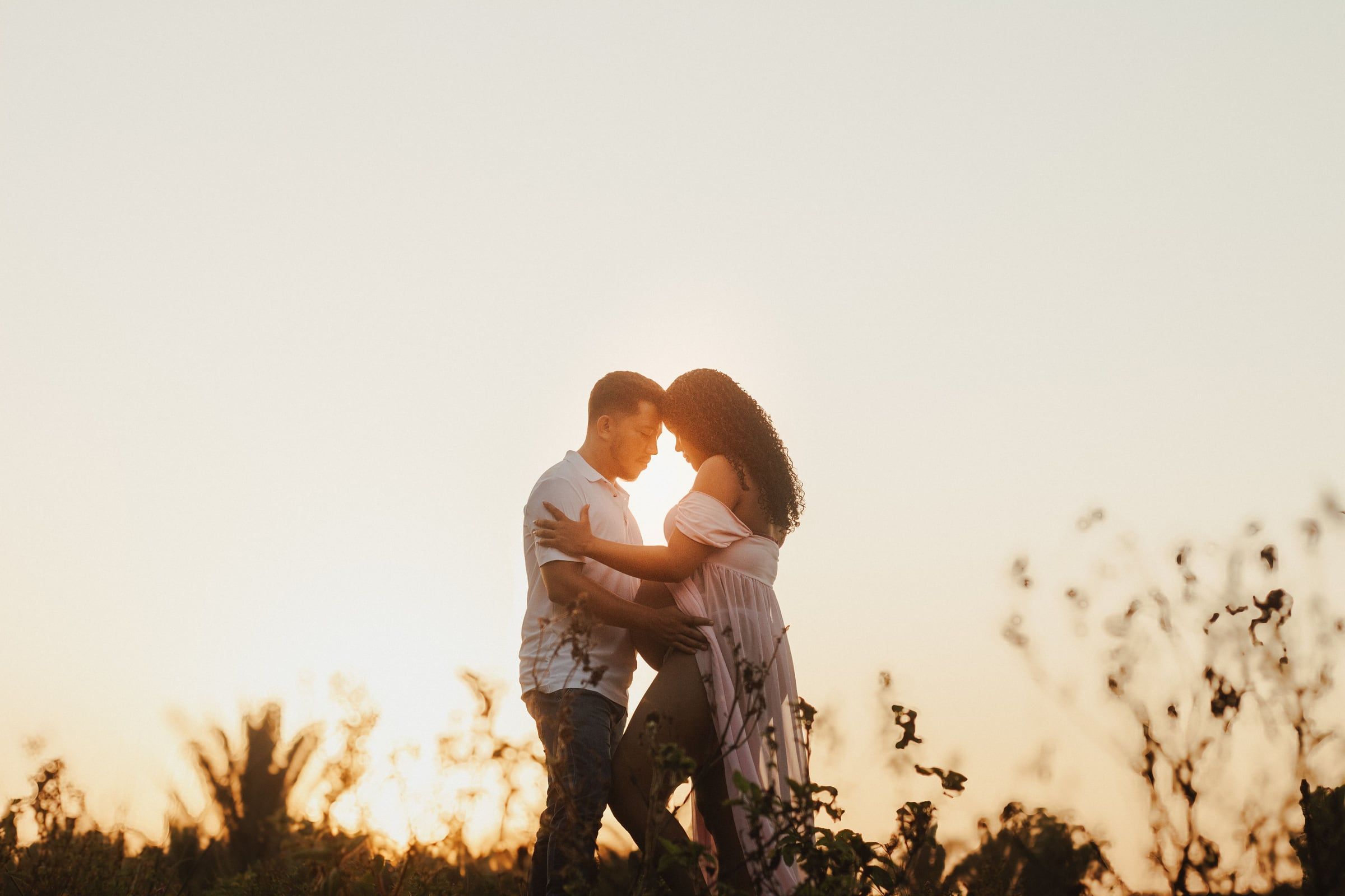 Casal abraçado em contraluz, ao pôr do sol, entre ervas altas.