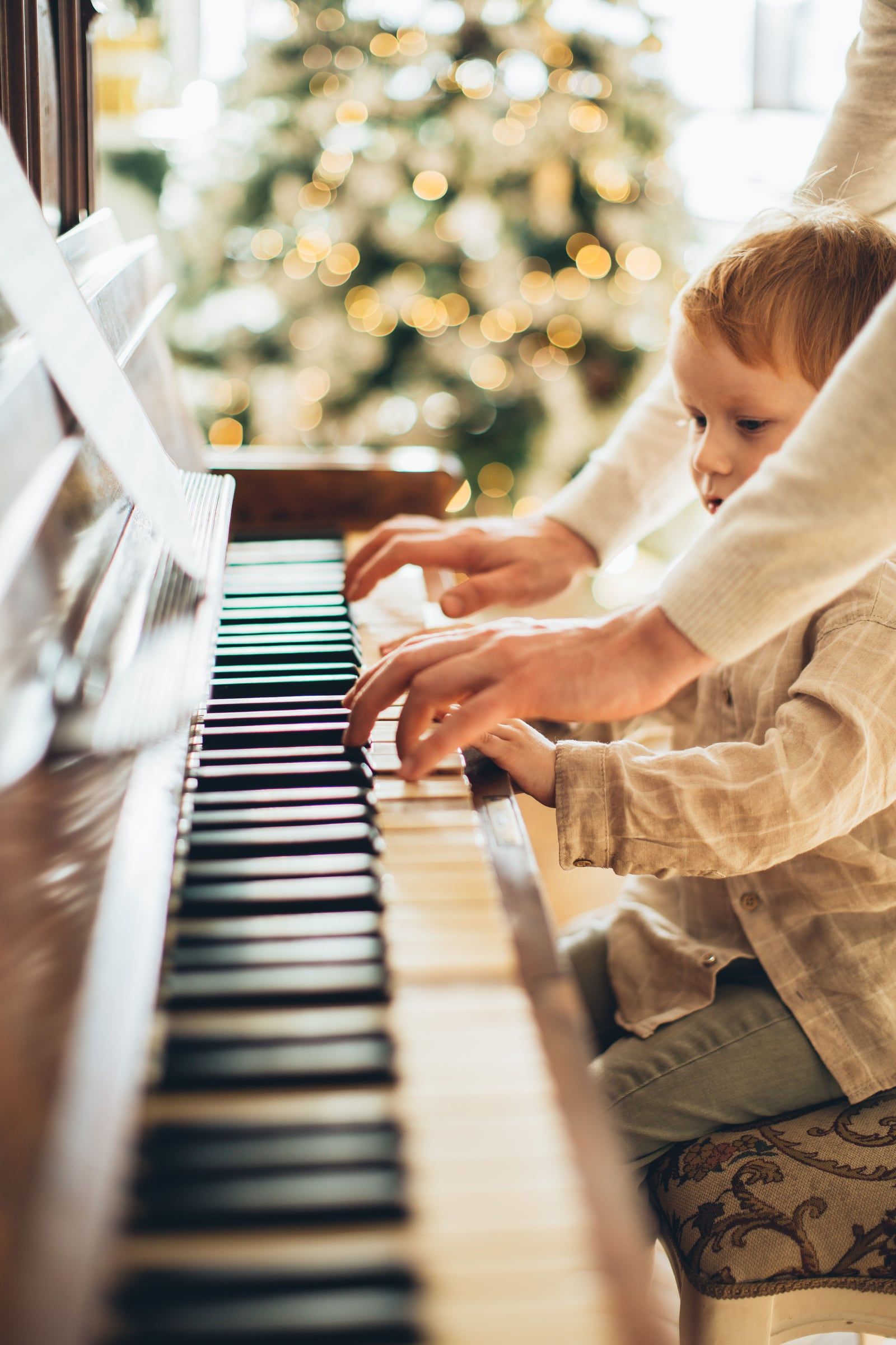 Mãos de um adulto e de uma criança no teclado de um piano, árvore de Natal ao fundo.