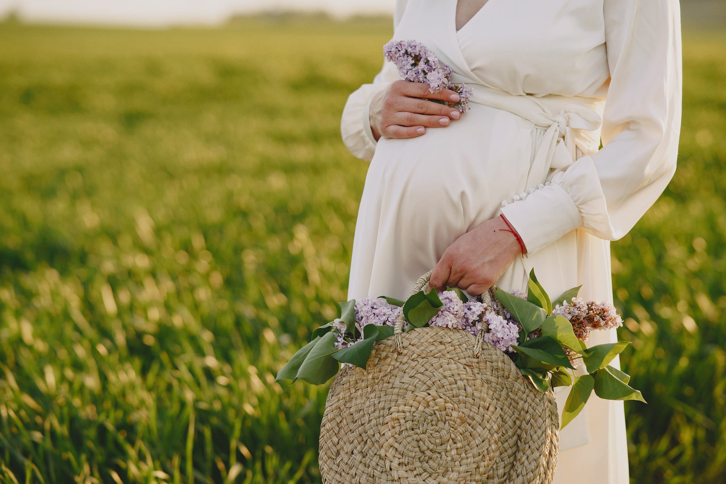 Barriga de grávida de vestido claro e um cesto de palha com flores lilás, num prado verde.