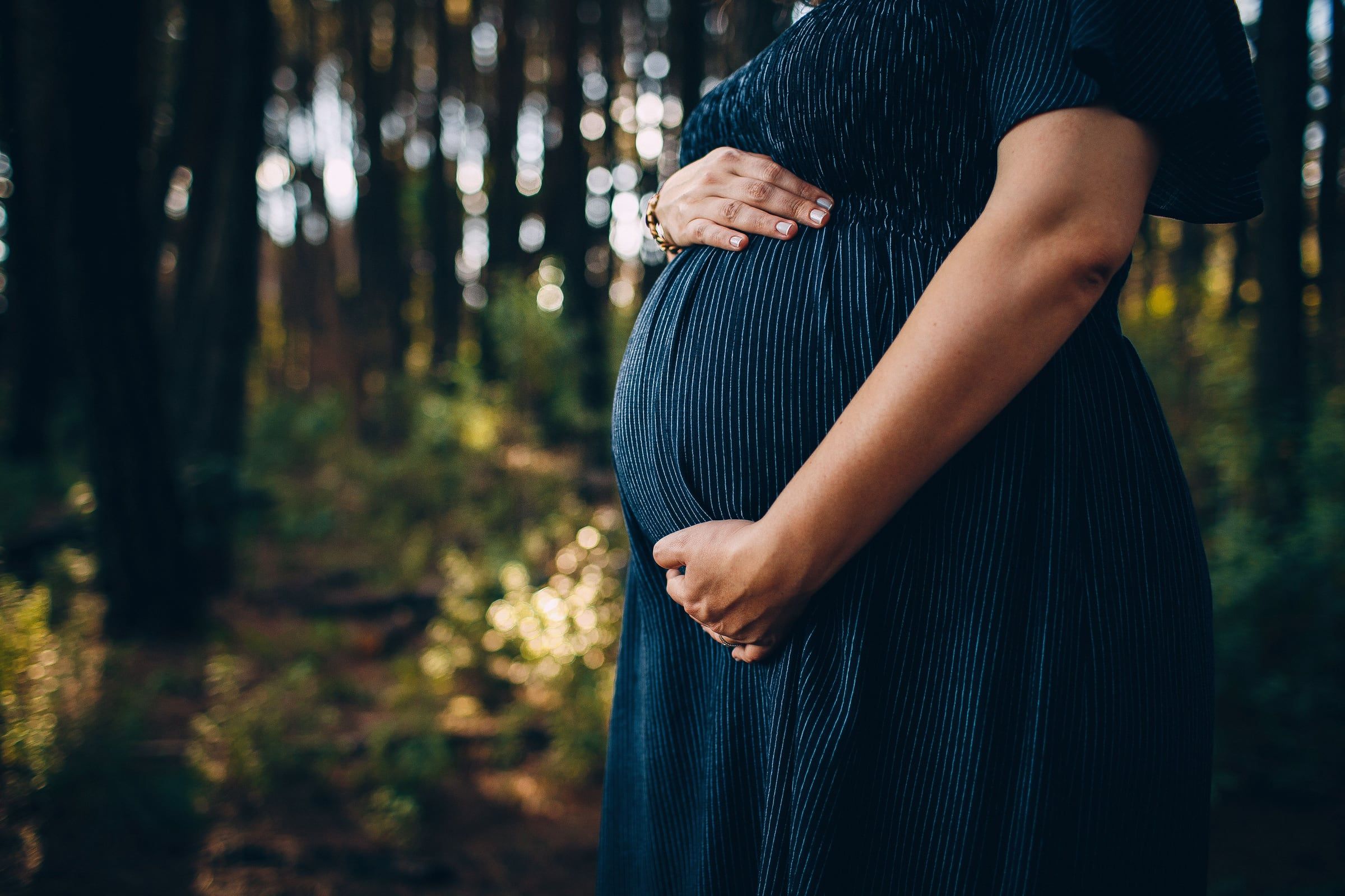 Mãos sobre a barriga, vestido azul escuro, pinhal ao fundo.