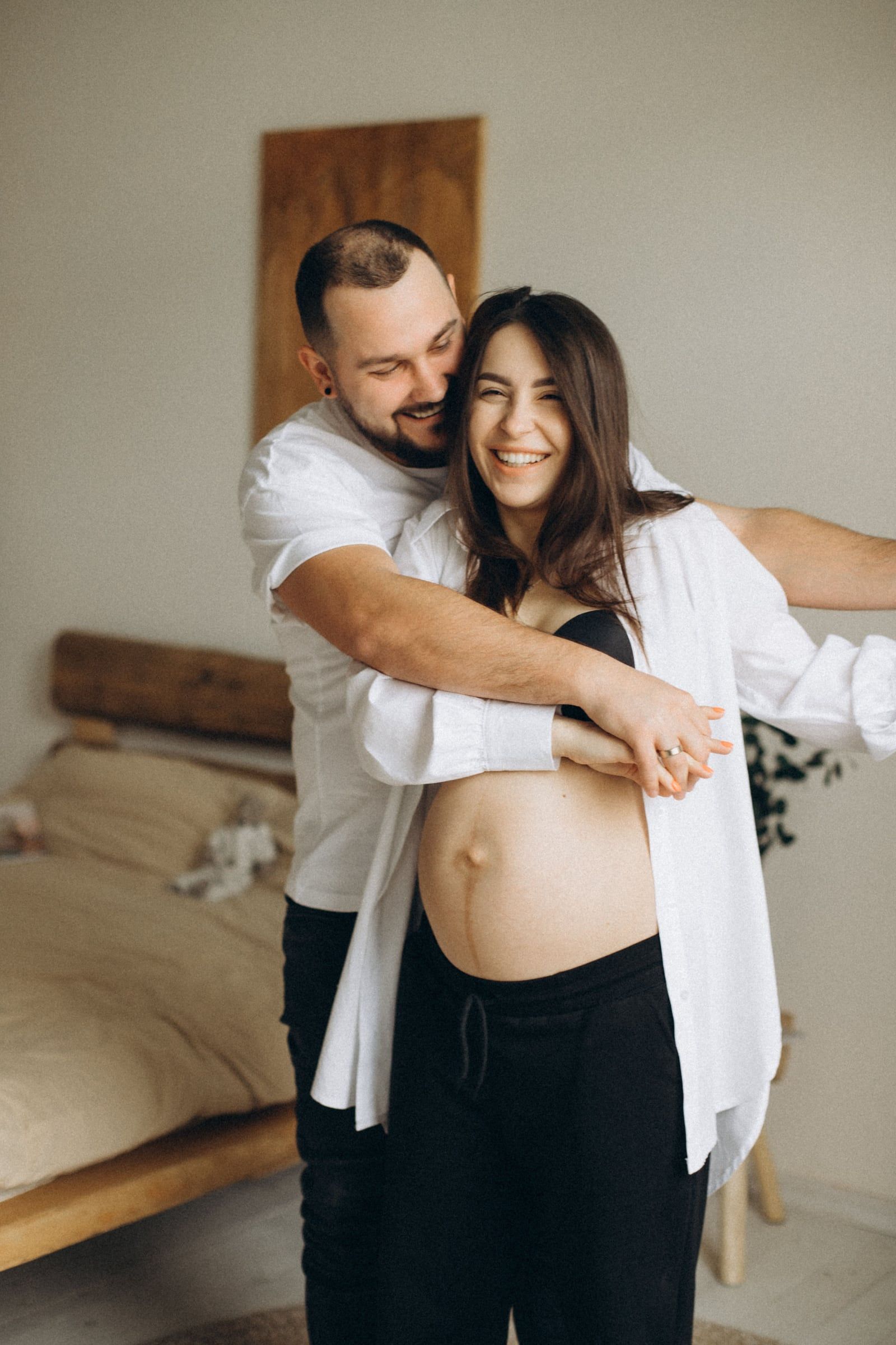 Casal a rir no quarto, ele abraça-a por trás, ela de camisa aberta sobre a barriga.