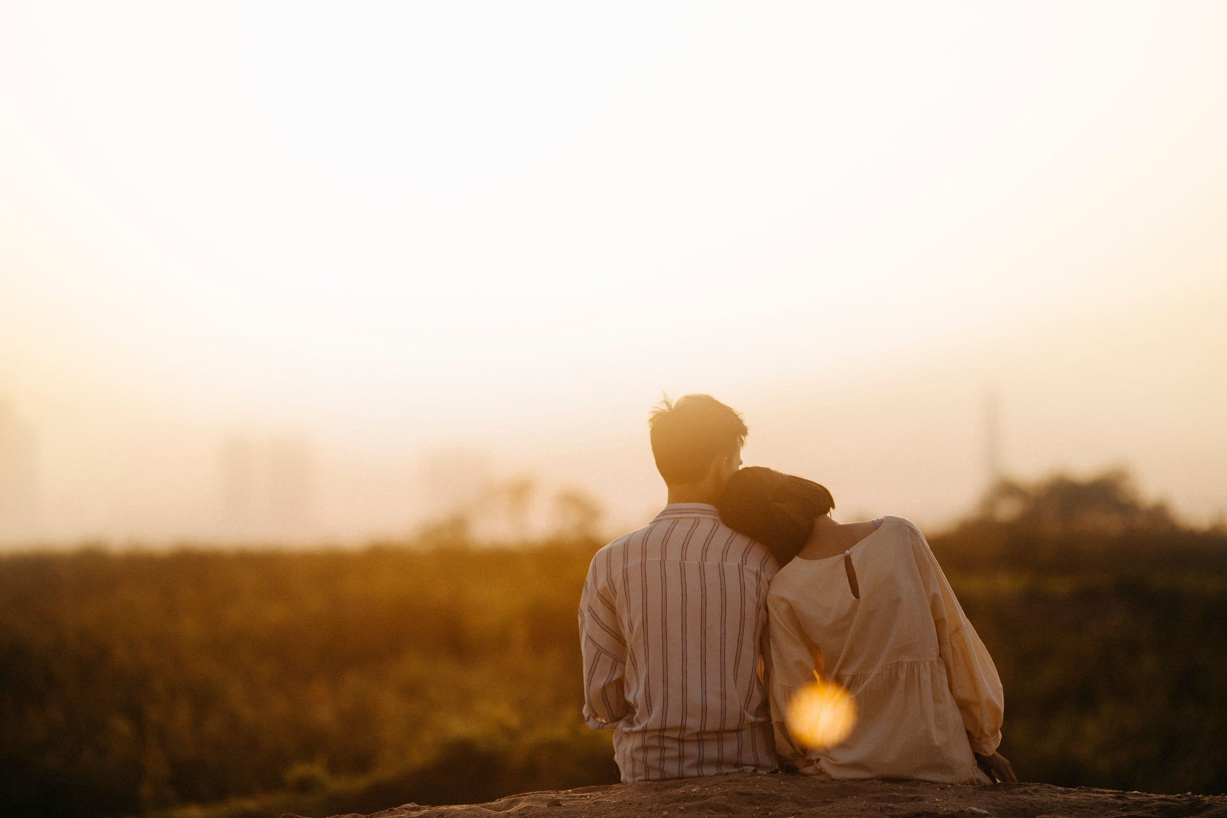 Casal sentado de costas ao pôr do sol, cabeças encostadas.