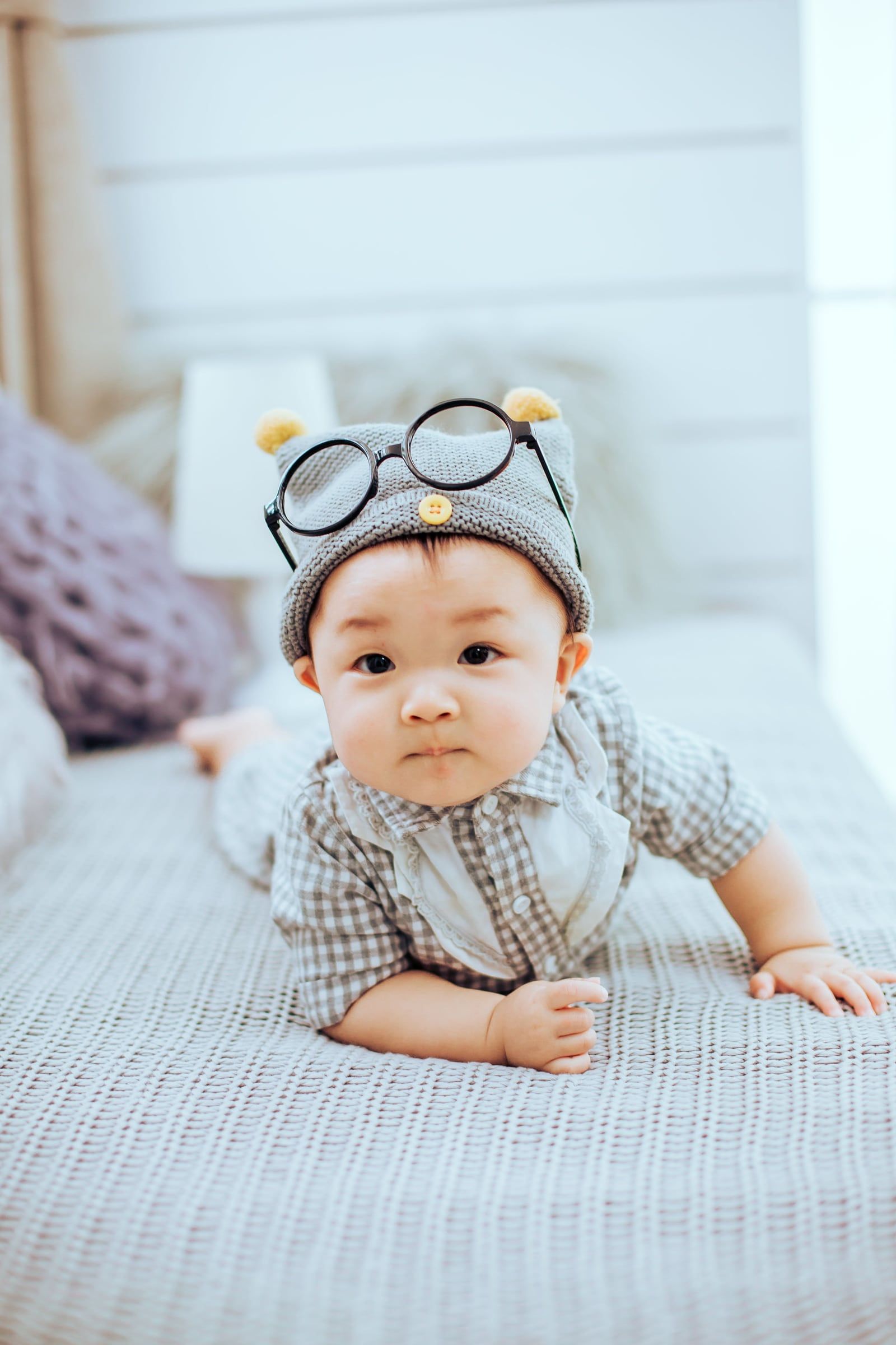 Bebé de gorro e óculos de brincar, deitado de barriga para baixo na cama.