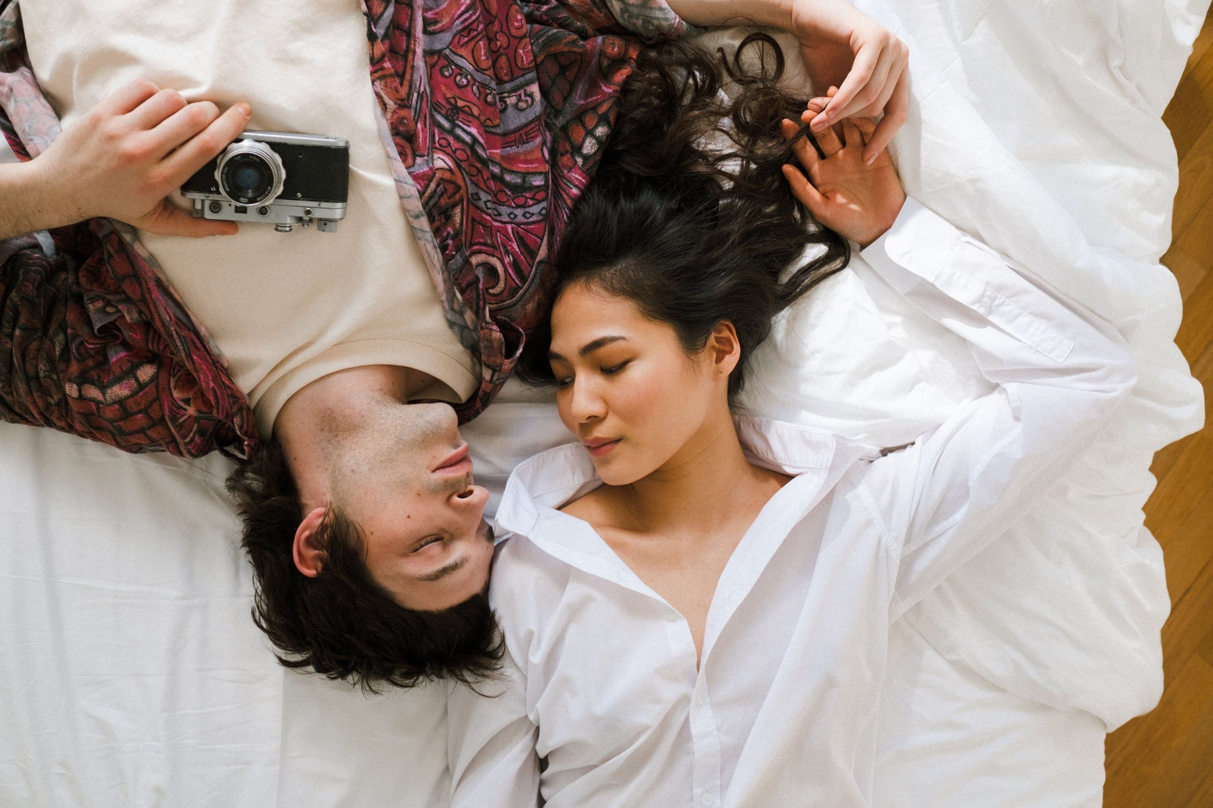 Casal deitado em lençóis brancos, cabeças juntas, uma câmara de filme ao lado.