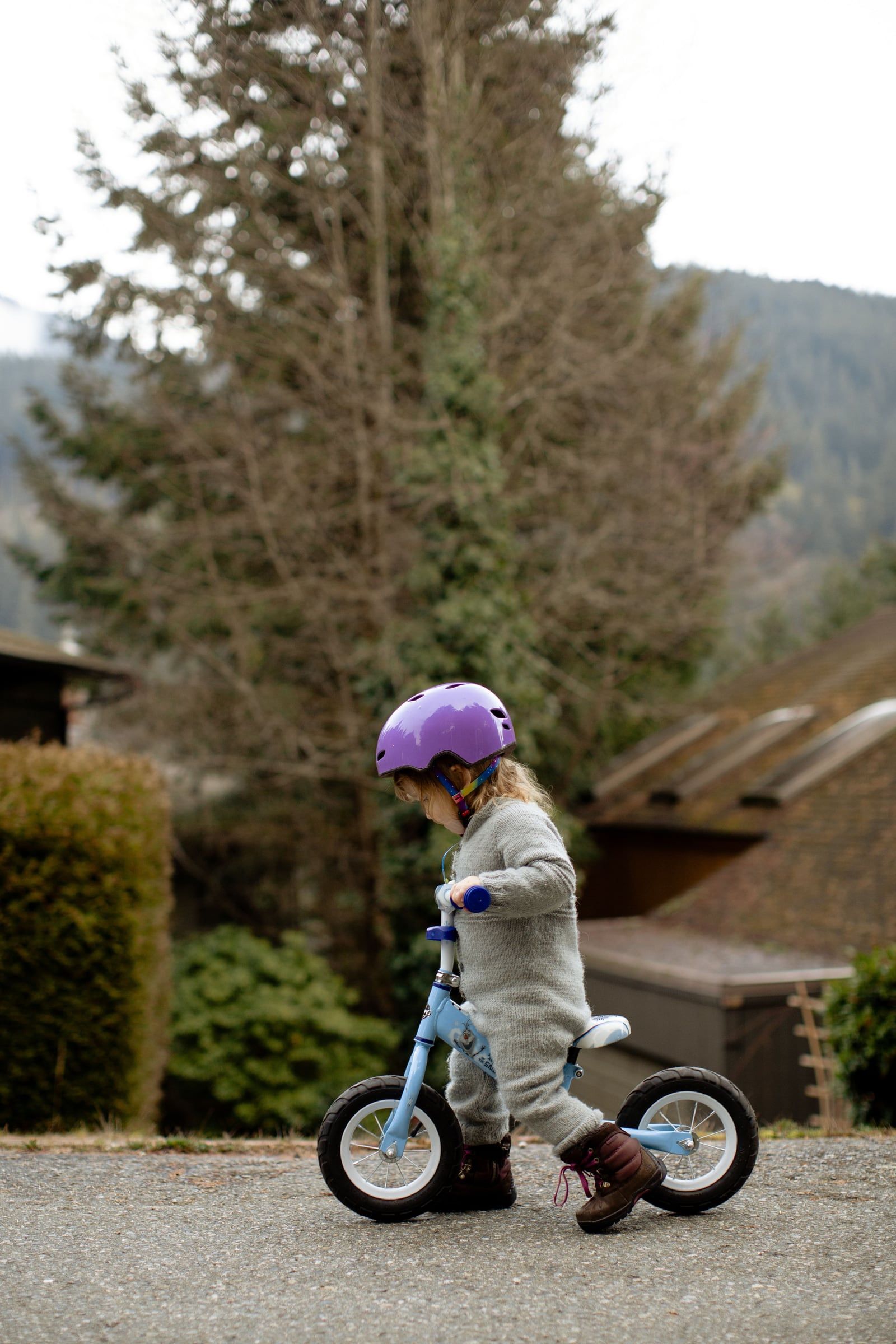 Criança de capacete lilás numa bicicleta sem pedais, em frente a uma casa.