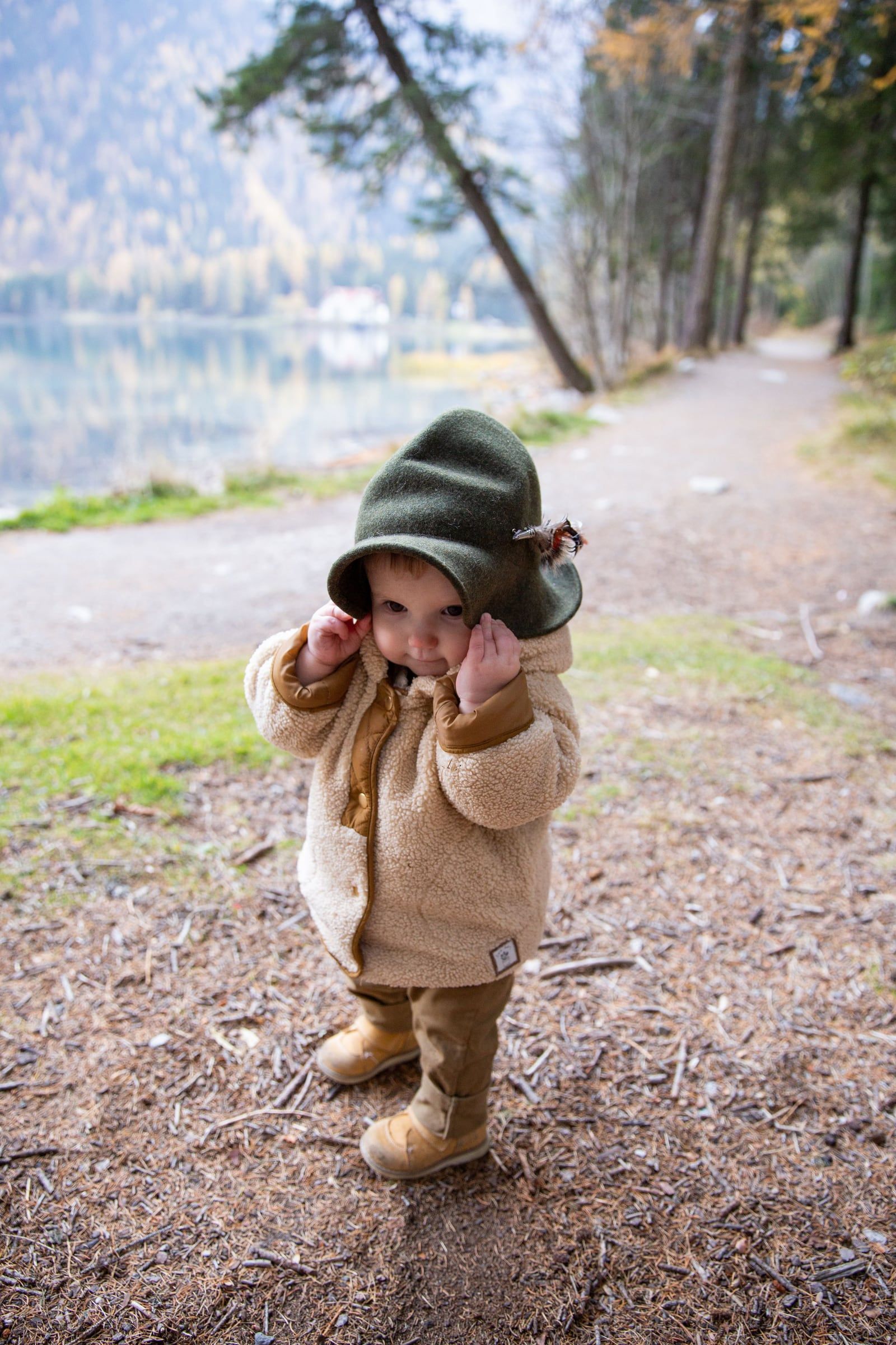 Criança pequena de gorro verde a esconder a cara, num caminho de terra junto a um lago.
