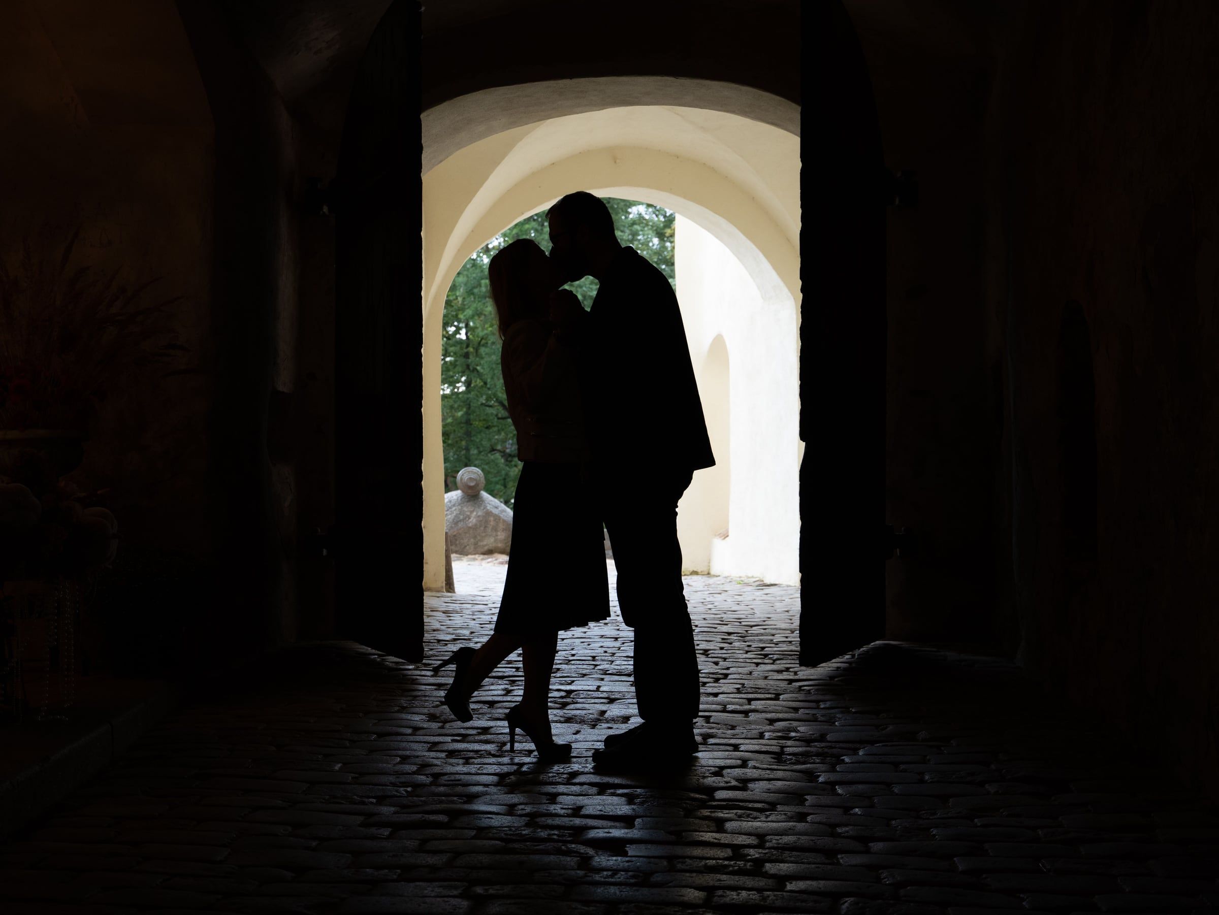 Silhueta de um casal a beijar-se debaixo de um arco de pedra, com luz ao fundo.